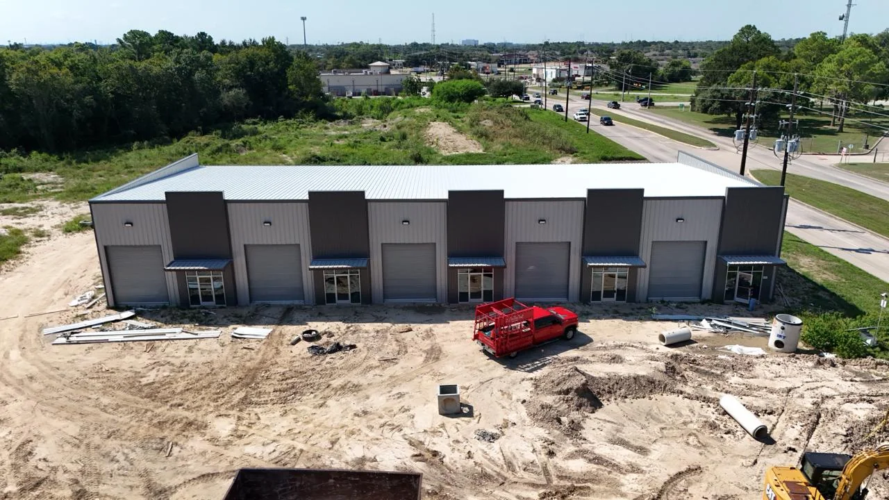 Aerial view of the Bammel front elevation with roll-up doors hung and storefront glazing installed across five tenant bays, apron still unpaved