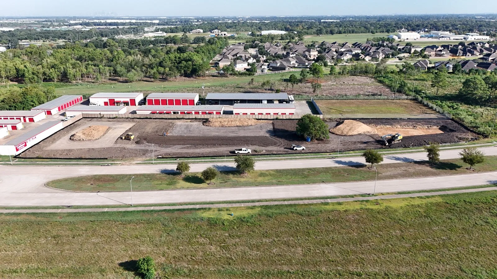 Aerial view of the cleared and graded McHard Flex lot with dirt stockpiles and equipment, next to existing storage buildings.